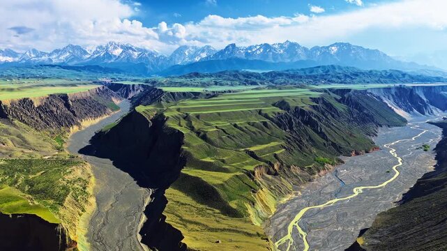 Aerial view of Anjihai Grand Canyon with dramatic green plateaus, winding riverbed, and snow-capped Tianshan mountains under a blue sky, Xinjiang, China.