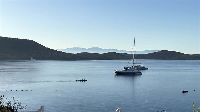 A wide, peaceful view of a calm blue bay featuring anchored luxury yachts, a rowing team in a scull boat, and scenic rolling hills beneath a clear morning sky.