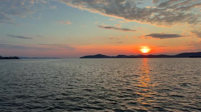 A calm, warm sunset over the Aegean Sea in Gulluk, Turkey. The sun hangs low in a hazy orange sky, casting a bright golden light path across the gently rippling water toward distant dark hills.