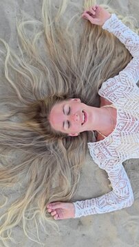 Young woman lying on sandy beach with eyes closed, long blonde hair spread out around her head in wide fan shape, wearing white lace top, top view