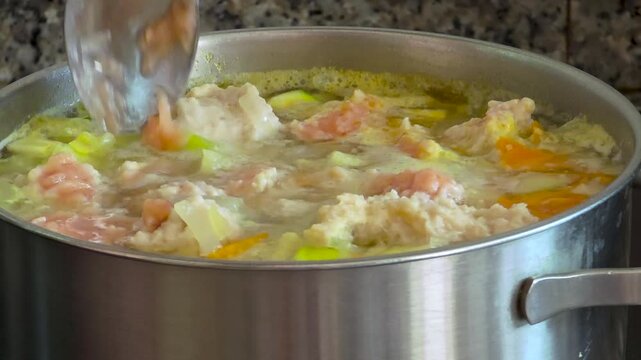 Slow motion close-up of adding raw minced meat dumplings into a boiling homemade vegetable soup with carrots and fresh dill in a stainless steel pot.