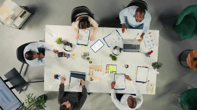 Close overhead framing follows diverse professionals in multinational office as they are collaborating on business planning, using performance metrics, report sharing and progress review.
