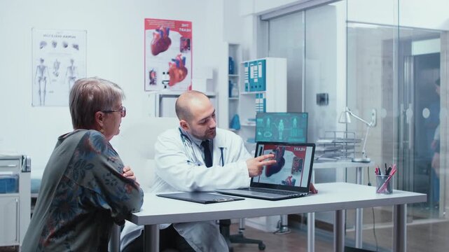 Young male medic presents a heart booklet on a computer to a senior woman patient, explaining cardiology risks in a private clinic.