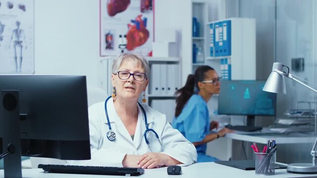 Experienced senior doctor smiles in a private hospital consultation room while a nurse works in the background, suggesting family therapy or patient care.