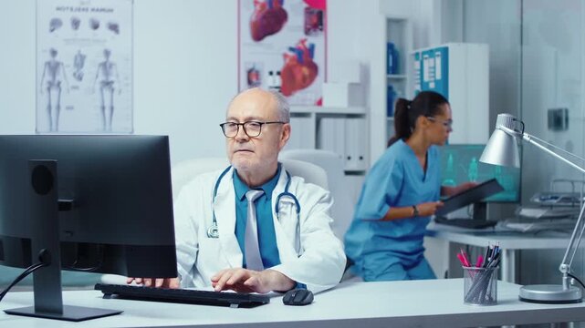 Senior doctor works at a computer and then smiles in a busy private clinic, connecting medical office workflow with professional healthcare staff.
