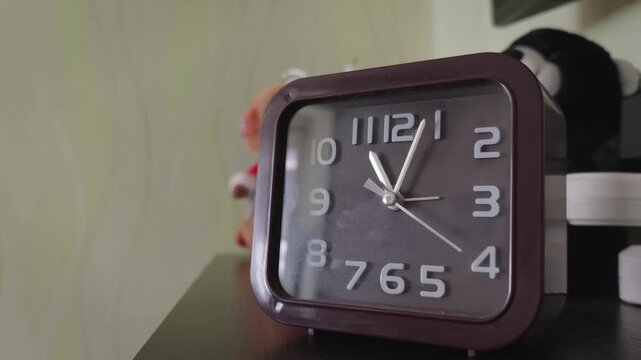 Close-up of a retro square analog clock with a dark face ticking on a wooden shelf. The minimalist interior setting suggests themes of time management and daily routines.