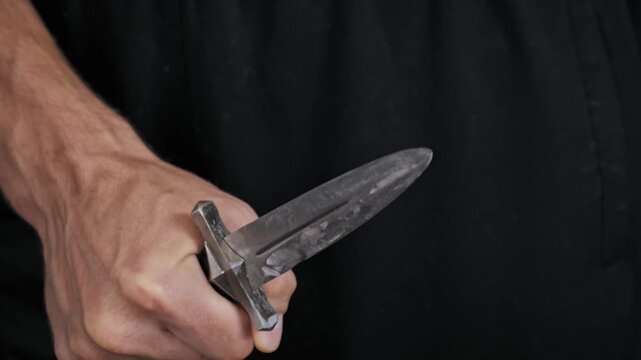 A close-up view captures a male hand tightly gripping a sharp double-edged dagger against a black background. Concept of criminal intent, danger, aggression, and threatening weapon possession