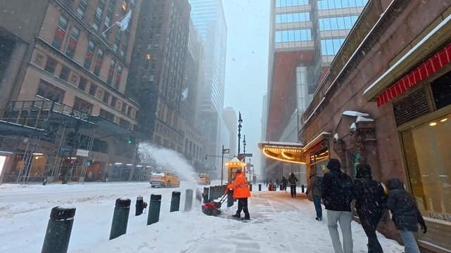 New York, USA, 25 December 2025 : Worker clearing snowy sidewalk with snowblower in New York. Person wearing orange safety jacket operating machine on snow covered urban street near building