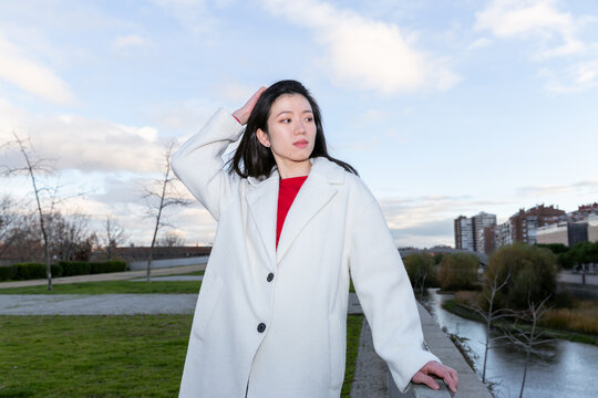Gen Z Asian woman in white coat by riverside at dusk