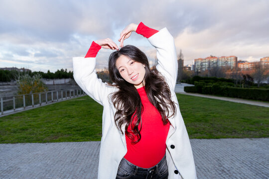 Gen Z Asian woman forming heart in city park at dusk