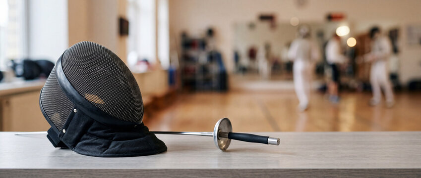Fencing mask and epee on bench in bright gym with athletes practicing in background