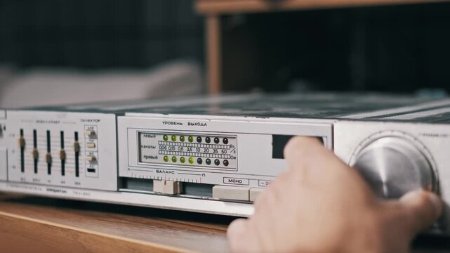 Side angle of a hand turning the silver volume knob on a retro stereo amplifier. The green LED level meter reacts to the audio signal change on the vintage hi-fi system.
