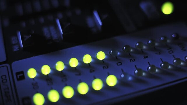Macro side angle of a sound mixer LED volume meter with yellow and orange lights pulsing. Faders are blurred in the background of a dark, blue-lit recording studio.
