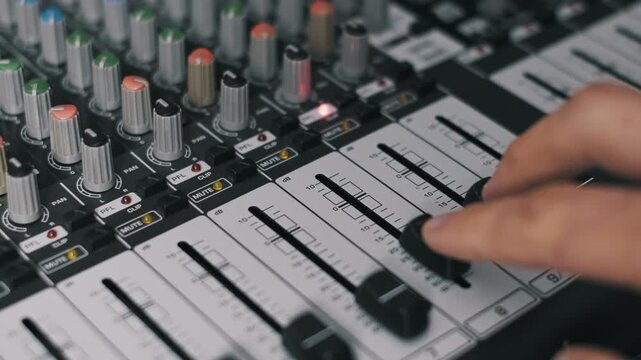 Macro of an audio producer's fingers moving a volume fader up and down on a mixing board. The shot reveals detailed decibel markings, various control knobs, and a red master fader.