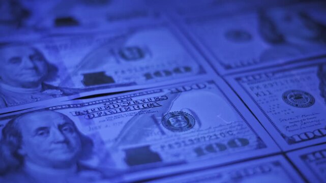 Close-up of hundred US dollar bills under shifting blue ultraviolet light slowly revealing banknote details. Shallow focus highlights paper texture and security features on a dark background.