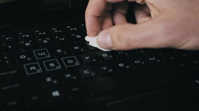 Close-up of a hand spraying antiseptic liquid on a black gaming laptop keyboard and wiping grime. The disinfection routine emphasizes health safety and maintenance of high-use electronics.