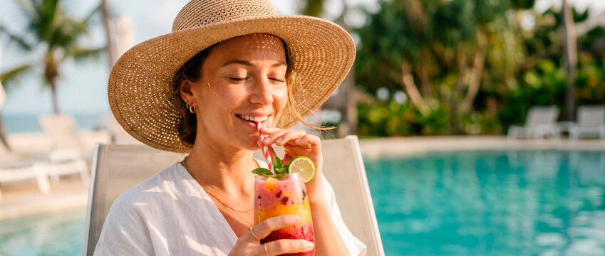 Woman in straw hat drinking tropical cocktail by pool. Luxury vacation resort lifestyle. Happy tourist relaxing at sunny summer beach club destination. Refreshing beverage and leisure.