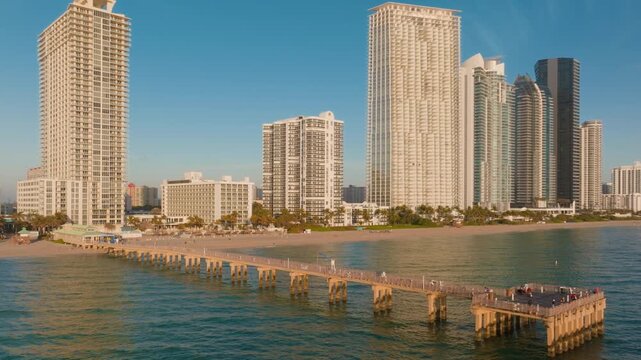 Aerial drone footage of Sunny Isles Beach fishing pier extending into turquoise Atlantic Ocean waters with luxury highrise condominiums and beachfront hotels lining the sandy Miami coastline.