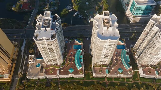 Stunning aerial drone footage captures luxury high-rise condominium towers in Sunny Isles Beach Miami during golden hour with rooftop pools and palm trees along the waterfront canal district.