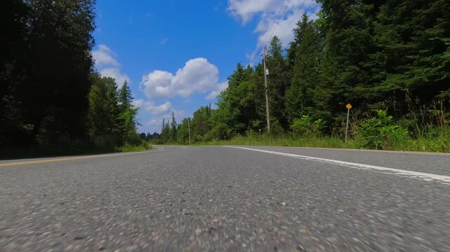 Rural countryside road drive with rolling hills, bright sunlight and cloudy blue sky. Gentle turns, captured from a steady front exterior view from below the bumper of the vehicle.