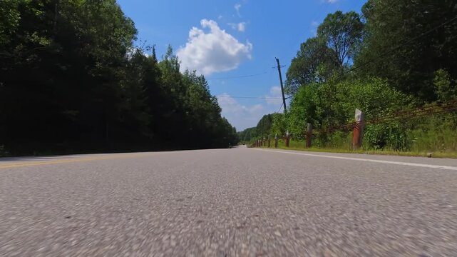 Rural countryside road drive with rolling hills, bright sunlight and cloudy blue sky. Gentle turns, captured from a steady front exterior view from below the bumper of the vehicle.