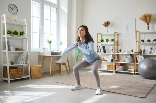 Bodyweight squats and sports exercises by happy woman in living room. Home cardio workout on yoga mat of girl in sportswear, athlete training body strength and squatting. Slimming, fitness concept