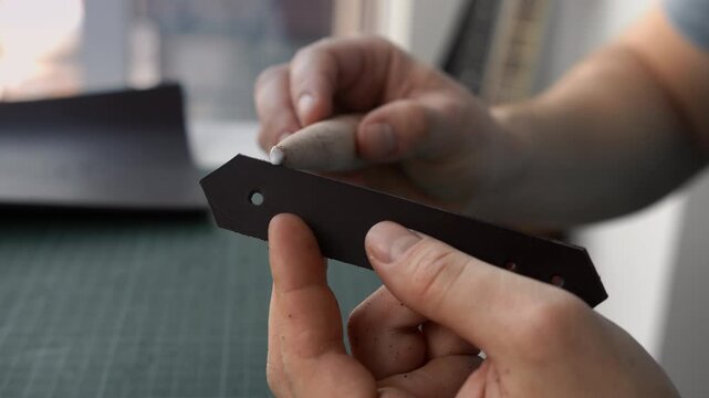 Leather craftsman burnishing a brown strap edge with a wooden slicker tool in a workshop creating handmade goods