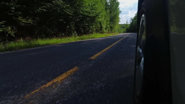 Rural countryside highway asphalt road drive. Cinematic POV of the front left car wheel driving plate. Yellow line lane separator in the frame. Cottage country commute, back of wheel exterior view.