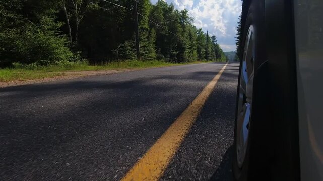 Rural countryside highway asphalt road drive. Cinematic POV of the front left car wheel driving plate. Yellow line lane separator in the frame. Cottage country commute, back of wheel exterior view.