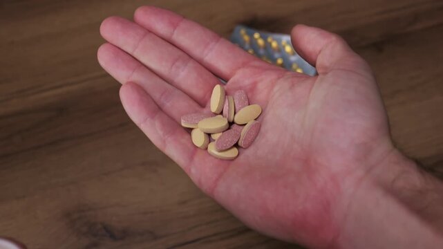 A close-up of a person pouring prescription medicine tablets from a white plastic bottle into their open palm. Blister packs of yellow capsules rest on the wooden table in the background.