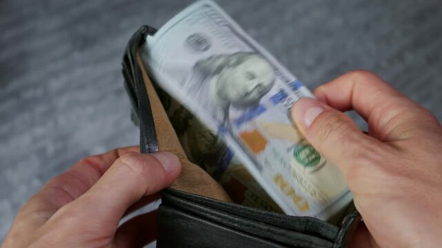Hands open a black leather wallet stuffed with a thick stack of hundred dollar bills against a textured background. Concept of financial success, wealth, cash savings, and prosperity.
