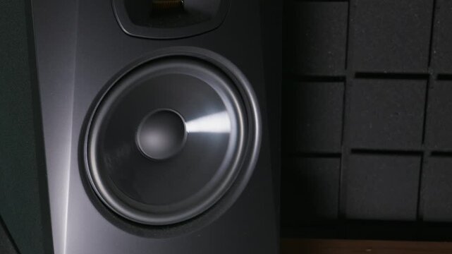 Close-up view of a black professional studio monitor speaker woofer vibrating with deep bass. The diaphragm moves rapidly against dark acoustic foam panels in a studio setup.