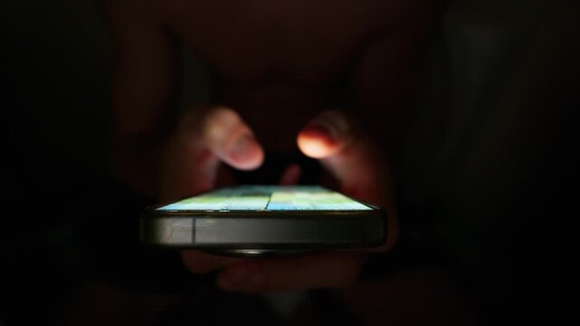 Male hands hold a glowing smartphone in a pitch black room. The bright screen illuminates his fingers as he scrolls through social media feeds. This shows modern digital addiction.
