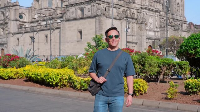Young smiling Latino man feeling confident, relaxed while walking forward looking around, enjoying a sunny day in Mexico City near a historic cathedral, wearing casual outfit with sunglasses and bag
