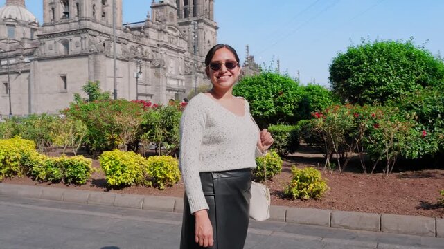 Young smiling Latina woman feeling joyful, relaxed and confident while walking and looking around, enjoying a sunny day in Mexico City near a historic cathedral, wearing casual chic outfit