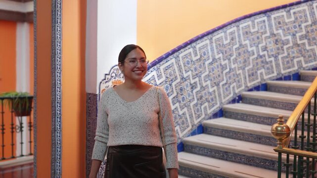 Young smiling Latina woman feeling happy, relaxed and confident while walking and looking around inside a hotel in Mexico City historic, surrounded by colorful tiled stairs, wearing casual chic outfit