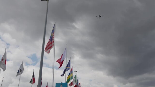 Low angle view of international flags waving on flagpoles with an airplane flying overhead against a cloudy sky