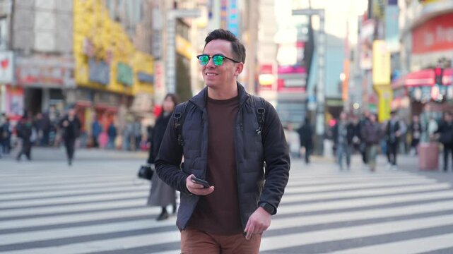 Young smiling Latino man feeling relaxed, happy and engaged while walking and checking his smartphone across Shibuya Crossing in Tokyo Japan, surrounded by busy streets crowds, wearing casual outfit