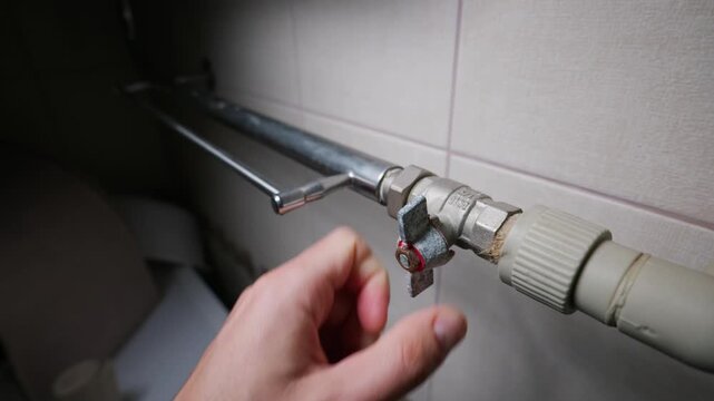 A male hand turns a metal ball valve handle to shut off the home heating or water supply system. Concept of home maintenance, plumbing repair, saving resources, and heating system control.
