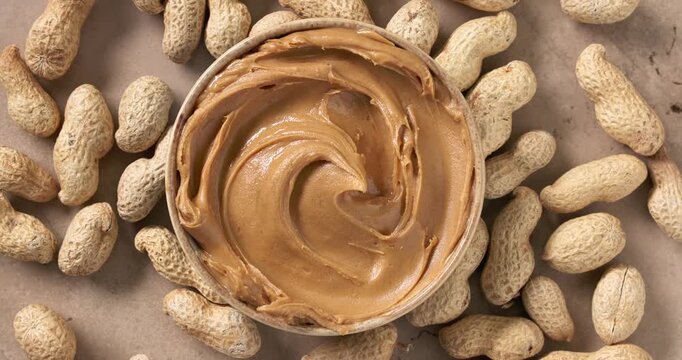 creamy peanut butter with a smooth decorative swirl on the surface, rotating on a rustic kitchen tabletop surrounded by scattered peanuts. Rich and delicious nut spread with a silky texture