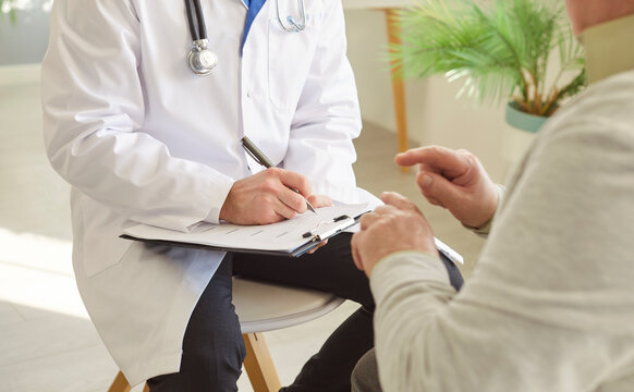 Cropped shot of faceless doctor cardiologist in white coat writing down prescription to old man patient, talking about ways of heart disease treatment, providing care. Medical appointment
