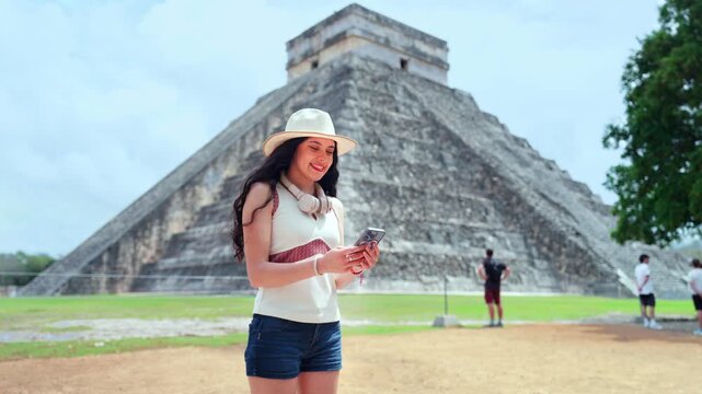 Young smiling Latina woman feels happy, curious while checking her smartphone at Chichen Itza in Yucatan Mexico, with Kukulcan pyramid ancient grounds behind her, wearing hat, sunglasses, casual top