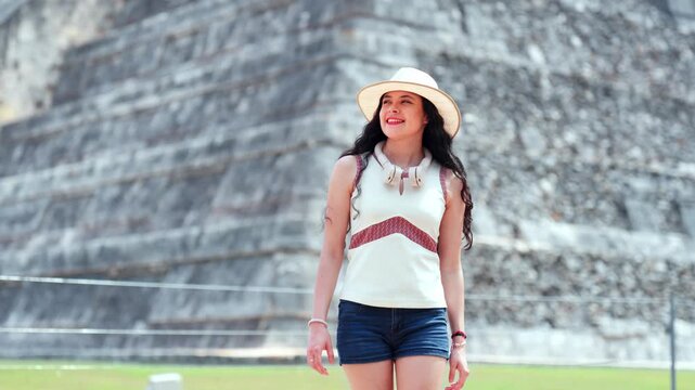 Young smiling Latina woman feels joyful, curious relaxed while walking and looking around at Chichen Itza in Yucatan Mexico, with Kukulcan pyramid behind her, wearing hat, sunglasses, casual outfit
