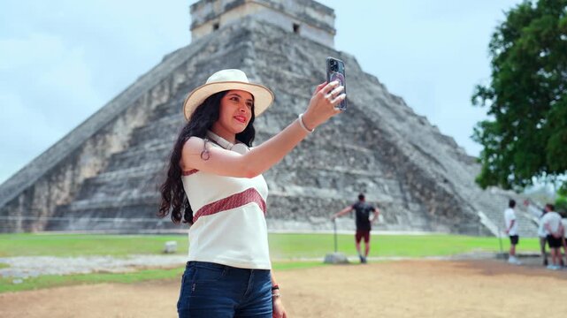 Young Latina woman smiling and taking a selfie with her smartphone at Chichen Itza in Yucatan Mexico, trendy travel moment at the iconic Kukulcan pyramid, enjoying sunny weather ancient Mayan ruins