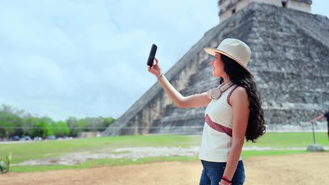 Young Latina woman smiling and taking a selfie with her smartphone at Chichen Itza in Yucatan Mexico, trendy travel moment at the iconic Kukulcan pyramid, enjoying sunny weather ancient Mayan ruins