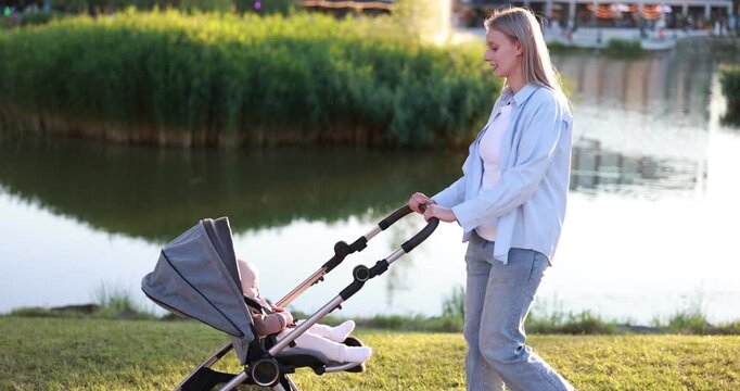 Mother walking with her cute baby in stroller at park on sunny day. Camera moving left