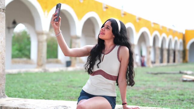 Young Latina woman smiling while taking a selfie with smartphone inside the iconic yellow convent of Izamal, Yucatan Mexico, surrounded by colonial arches, open courtyard, and historic architecture