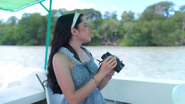 Young Latina woman on a small boat in a Yucatan lagoon using binoculars to search for flamingos, surrounded by calm water, mangroves, and rich wildlife during an ecotourism experience