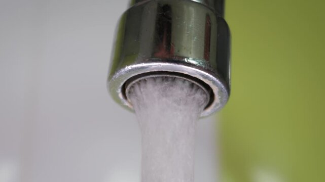 A chrome faucet pours a steady stream of aerated water into a sink. Metallic surfaces reflect bright lighting in a modern home. Soft green backgrounds highlight the clean flowing liquid.