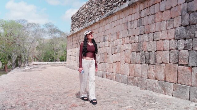 Young Latina woman exploring the archaeological site of Uxmal in Yucatan Mexico, walking next to an ancient Mayan stone wall, enjoying history, culture, and travel in a UNESCO heritage destination
